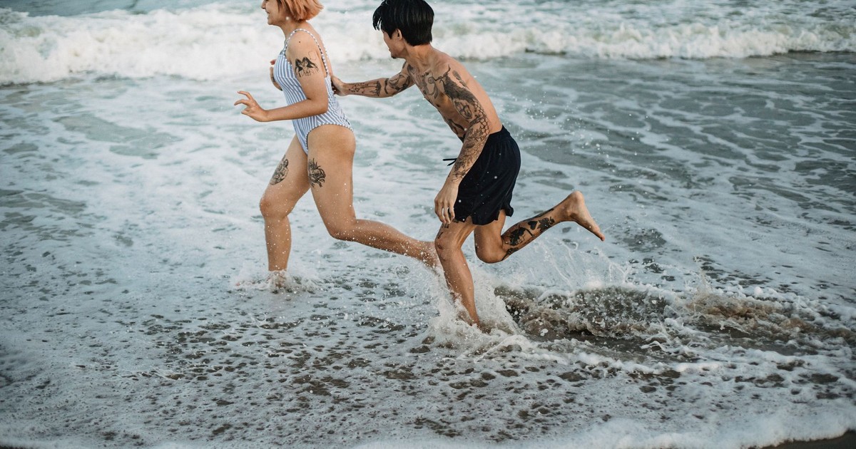 Tattooed couple having fun and running through the ocean waves on a sunny beach day.