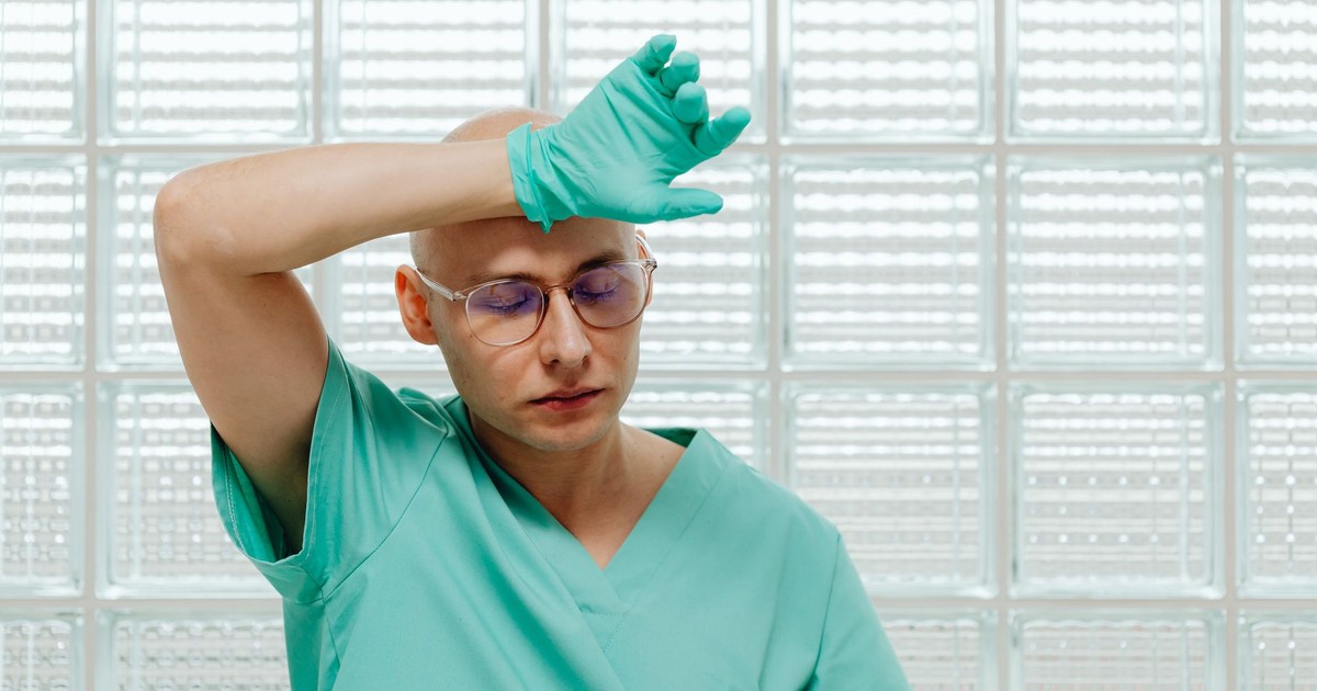 Tired male doctor wearing green scrubs and gloves, wiping brow in a medical setting.