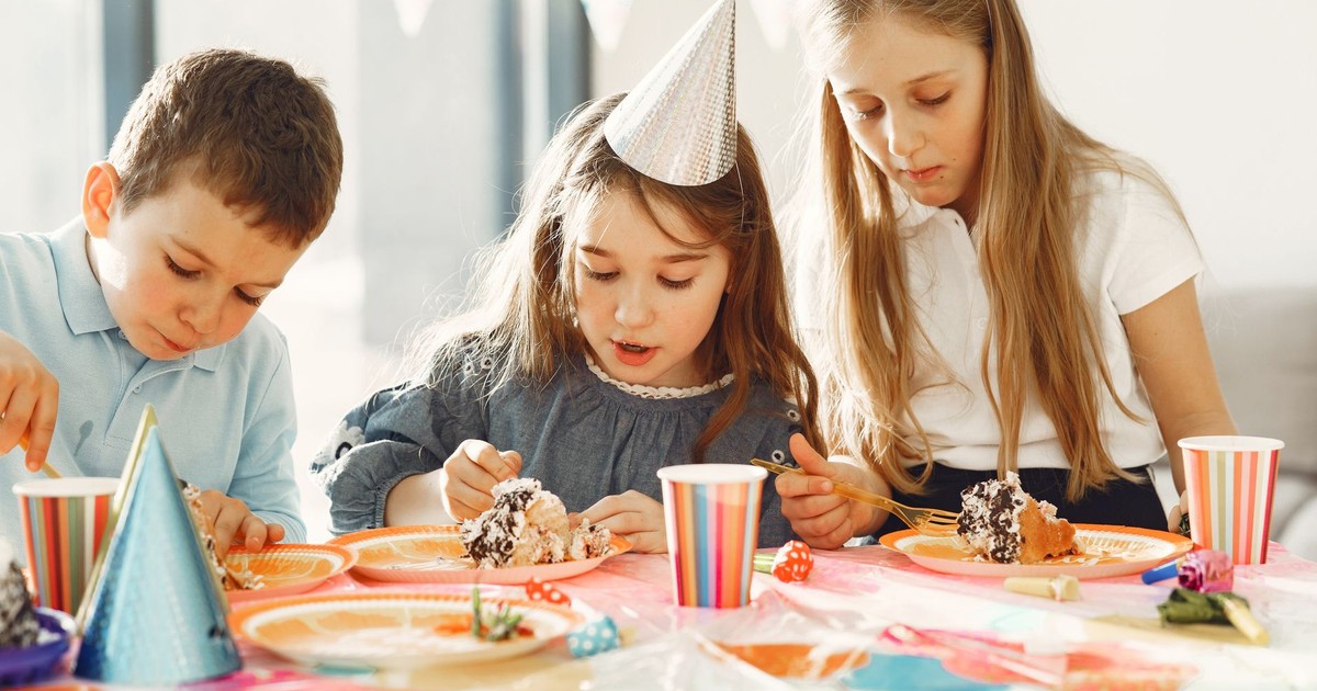 Three children enjoying cake at a colorful birthday party with hats and decorations.
