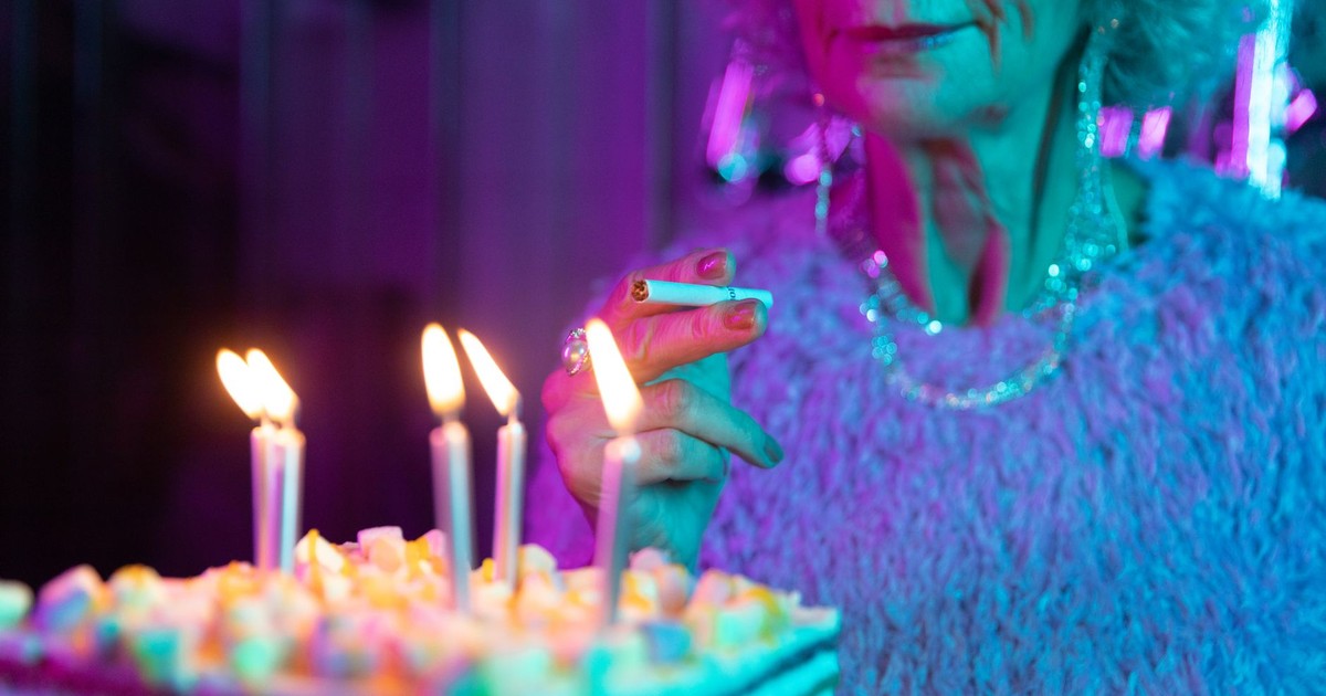 A senior woman holds a cigarette near a birthday cake with lit candles in a neon-lit room.