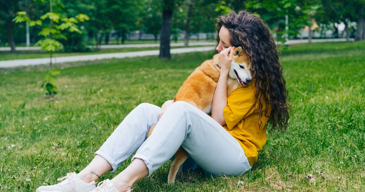 A woman sitting on grass hugging her Shiba Inu dog in a lush green park.