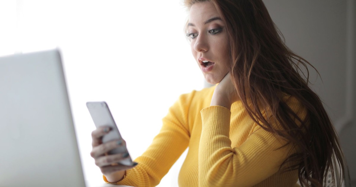Young woman in yellow sweater looks surprised at phone while working on a laptop indoors.