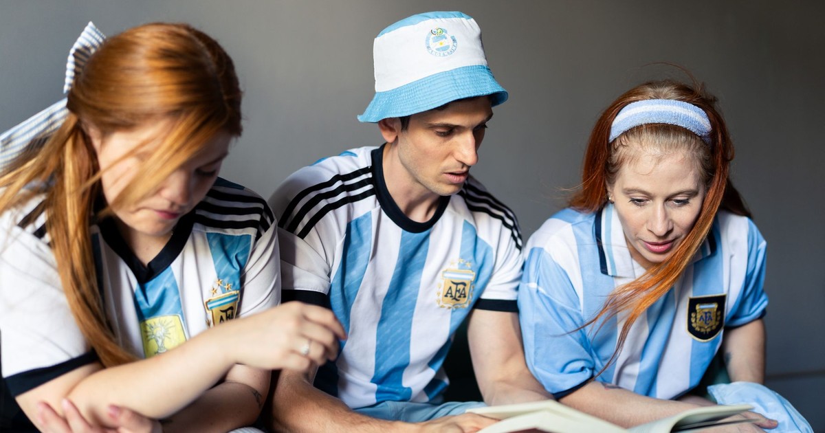 Three fans in Argentina jerseys engage with a book, showing team spirit indoors.