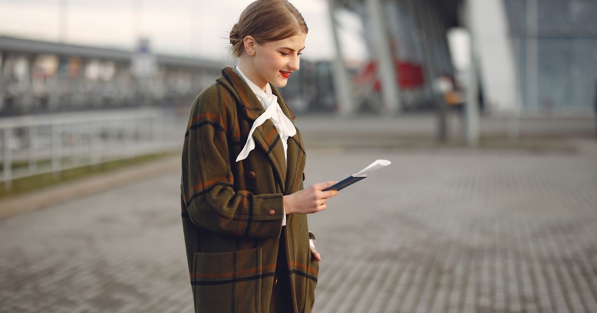 Smiling female passenger wearing trendy plaid coat and white blouse checking passport and ticket standing on pavement near contemporary building of airport
