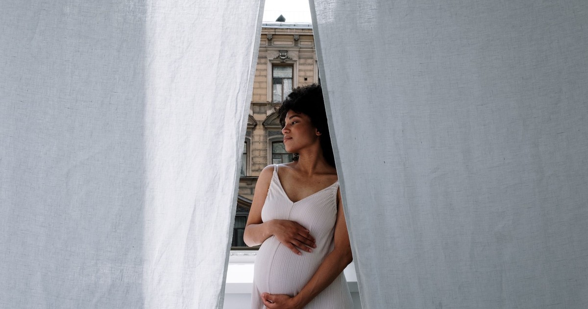 Elegant pregnant woman with Afro hair in a white dress standing thoughtfully by sheer curtains.