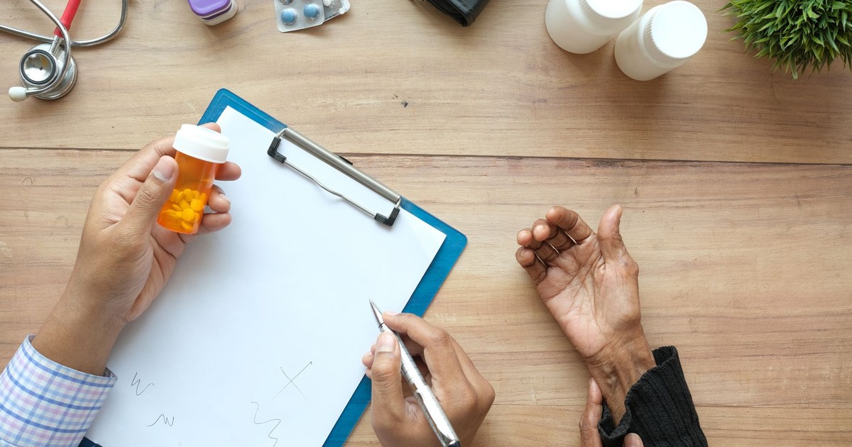 A doctor holds a pill bottle while consulting with an elderly patient. A stethoscope and prescriptions are visible.