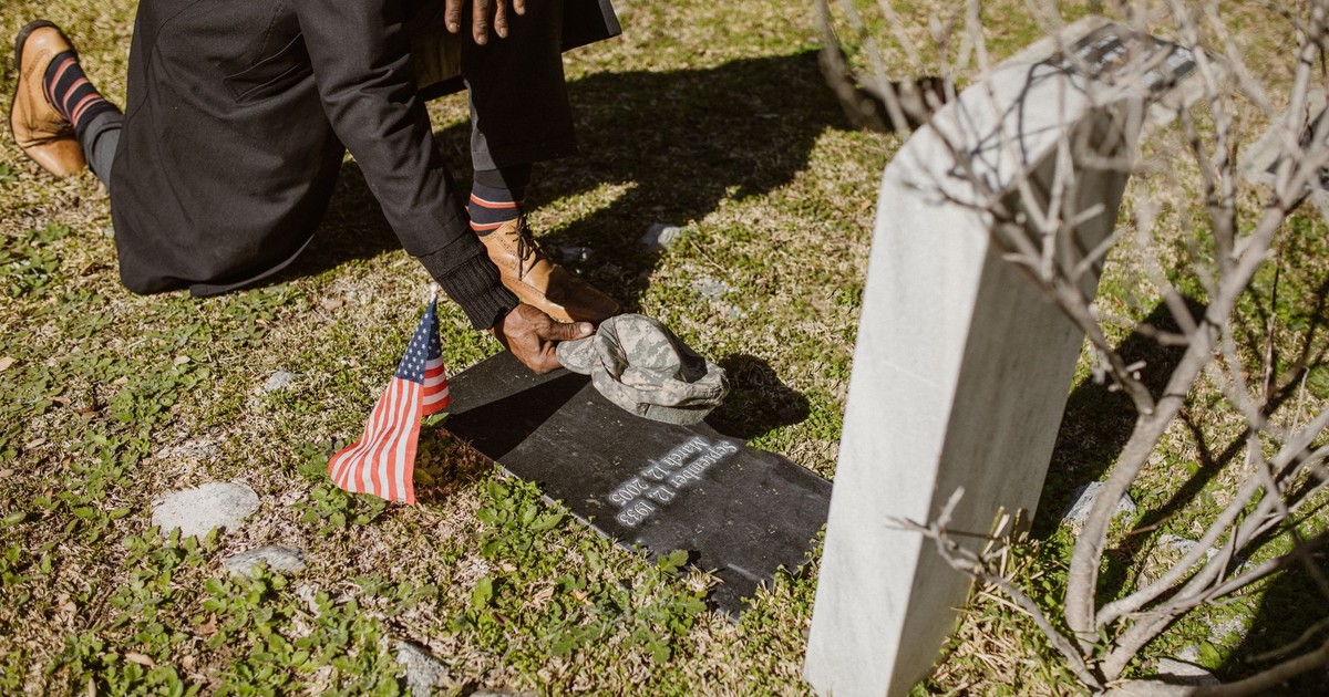 A person kneels by a gravestone with an American flag, paying respects at a cemetery.