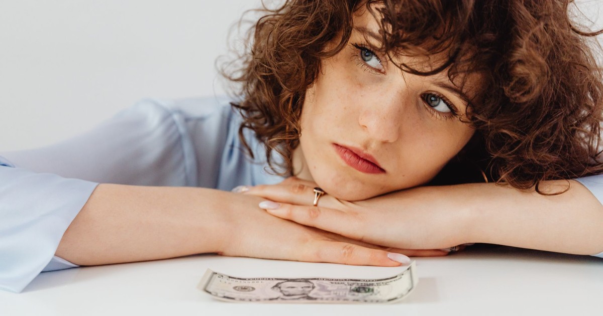 Worried woman with curly hair leaning on white surface with a US dollar bill.
