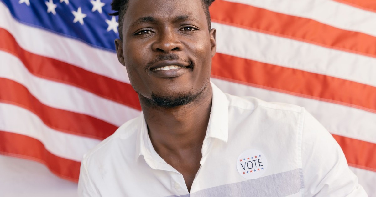 Adult man smiling with an 'I Voted' sticker and American flag backdrop.