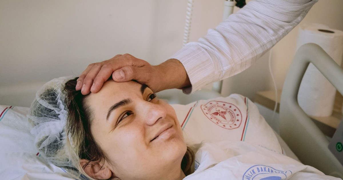 Smiling woman in hospital bed with supportive hand on forehead, conveying warmth and care.
