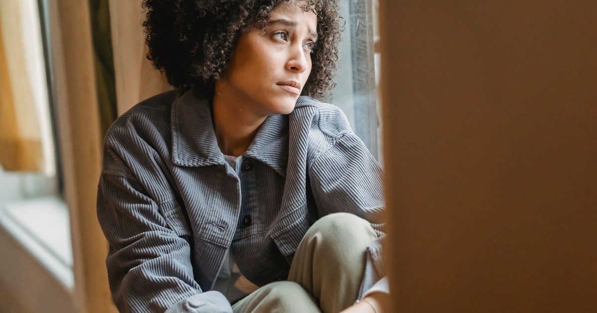 Frowning upset African American female with curly hair embracing knees while sitting on windowsill