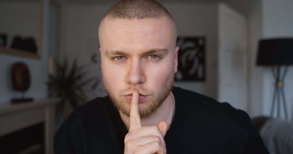 Portrait of a bald man with beard in a room, gesturing for silence. Mysterious expression.