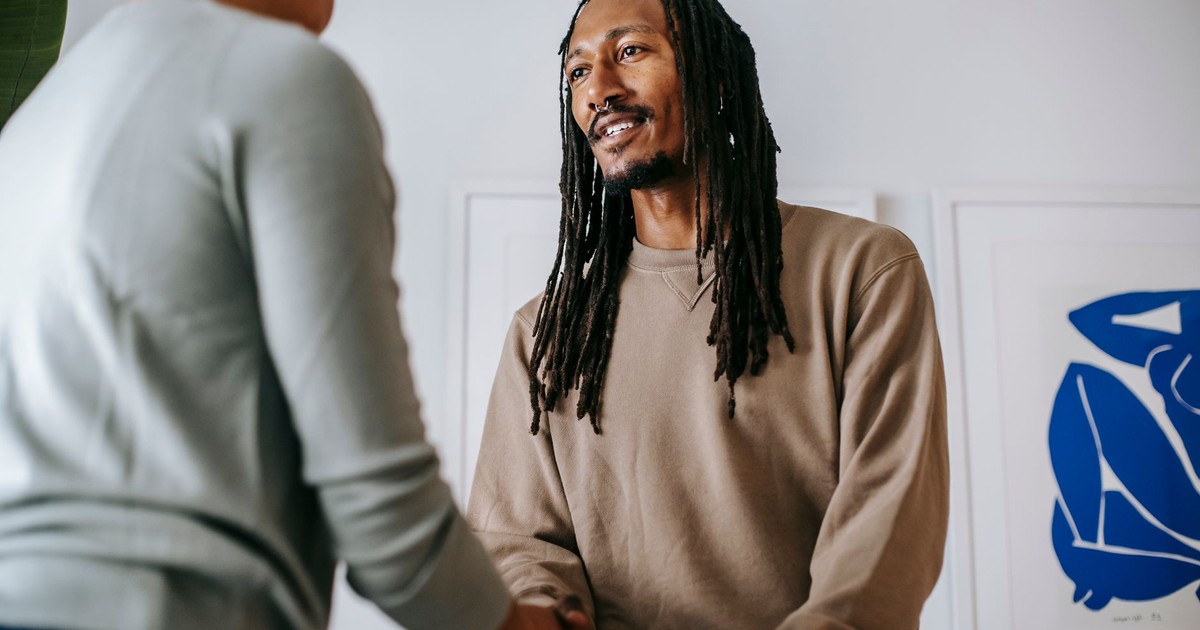From below of cheerful bearded African American male patient with dreadlocks shaking hand of unrecognizable female therapist while standing in office during appointment