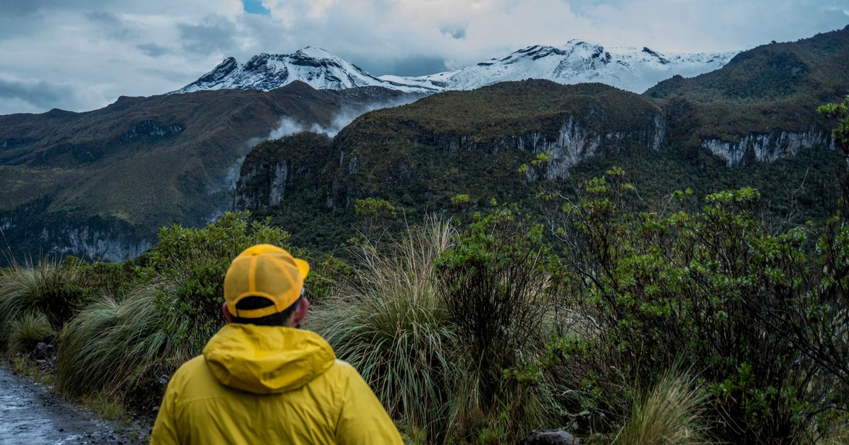 A man in a yellow jacket hikes through a scenic mountain landscape with snow-capped peaks.