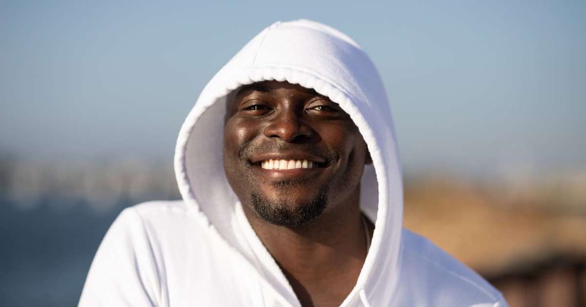 Close-up portrait of a smiling man in a white hoodie with a blurred outdoor background.