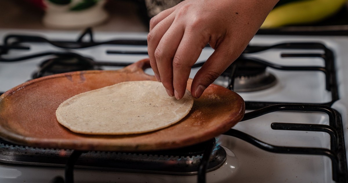 A hand placing a tortilla on a clay comal on a stove, preparing traditional Mexican food.