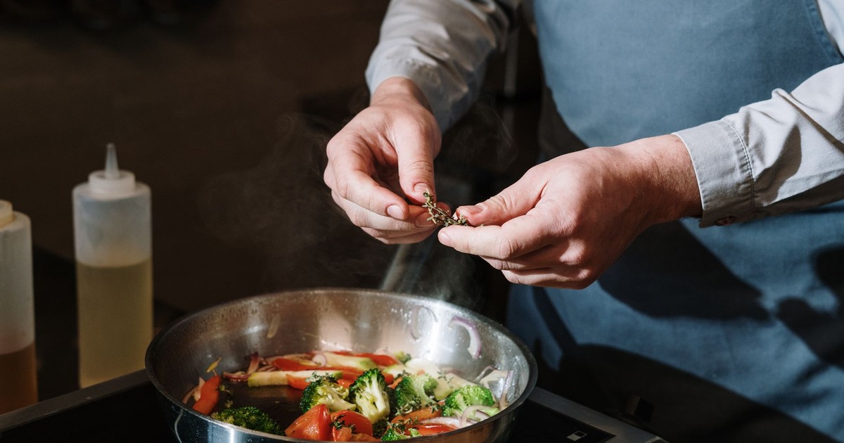 Chef seasoning a vegetable stir fry in a pan with fresh herbs on a stove.