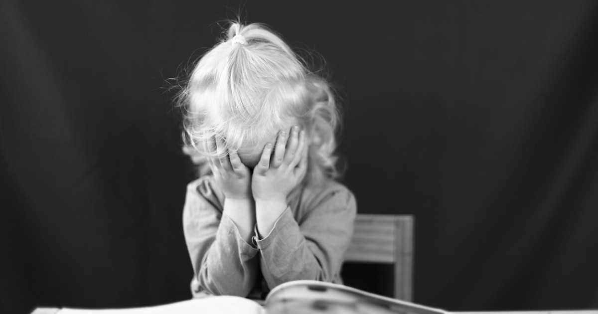 Black and white photo of a young child covering their face with hands, sitting in front of a book.