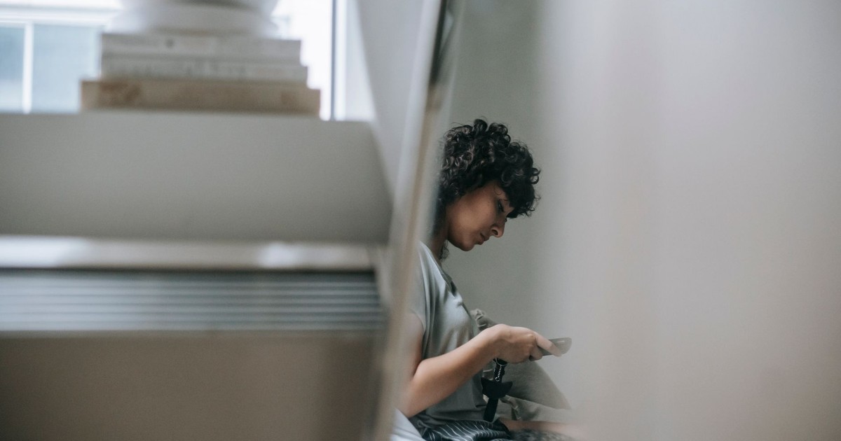 Side view of crop female text messaging on cellphone while resting near pile of books in house