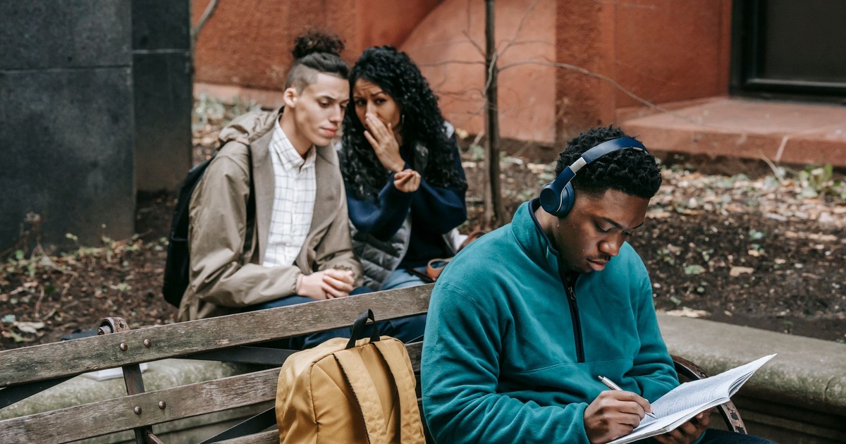 Diverse friends spreading gossip while sitting behind man with headphones and notepad on bench