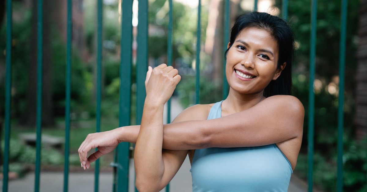 A woman in fitness attire stretching her arms outdoors with a confident smile.