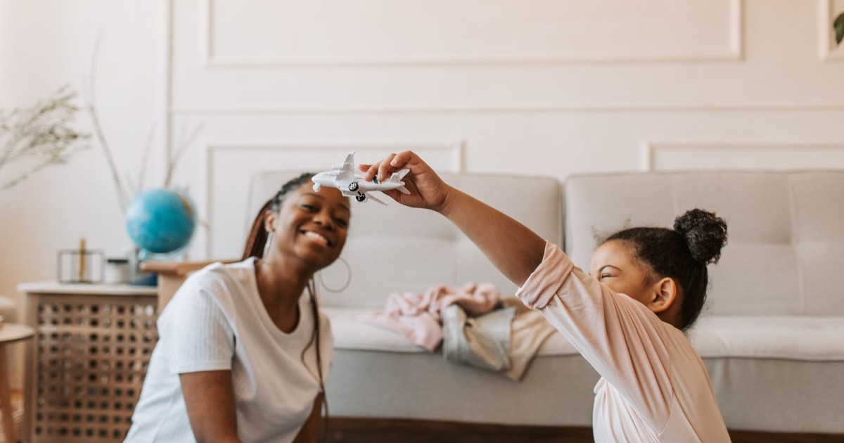 A young girl plays with a toy airplane while sitting with her mother on the floor indoors.