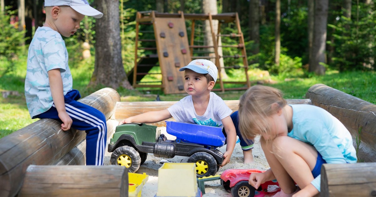 Three children engaged in imaginative play in an outdoor sandbox with toys.
