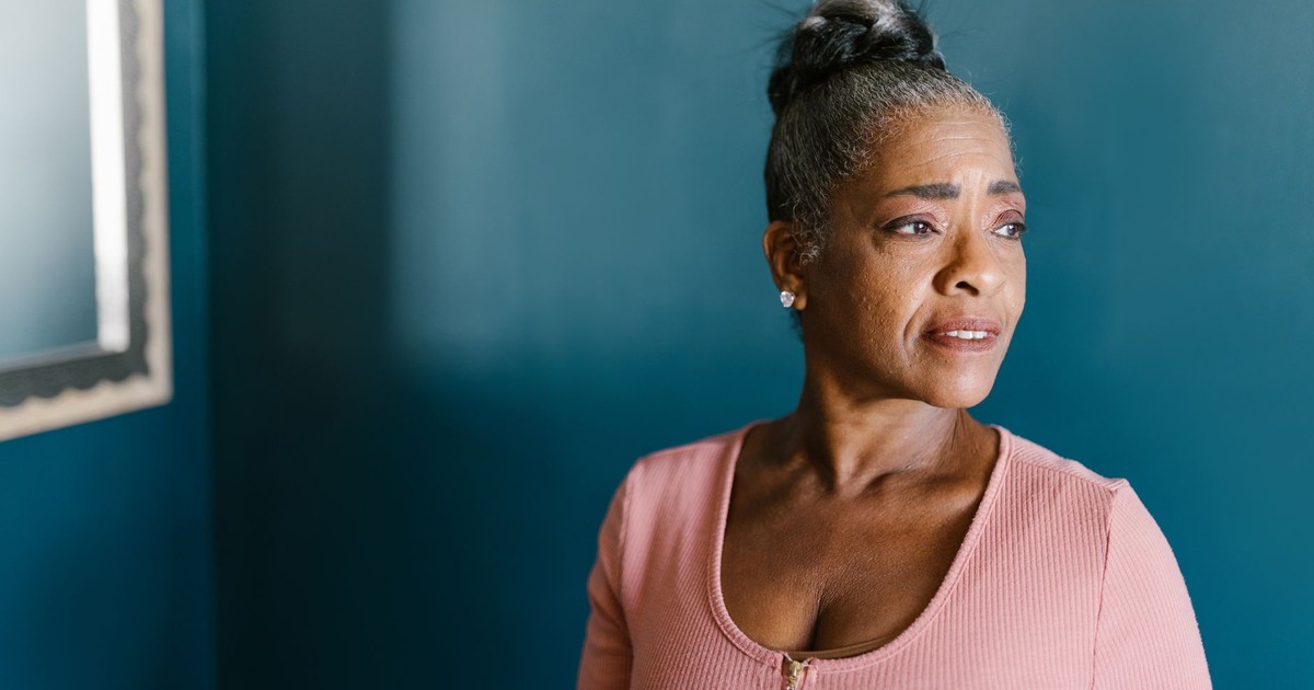 Elderly woman in pink gazing thoughtfully by a blue wall indoors.