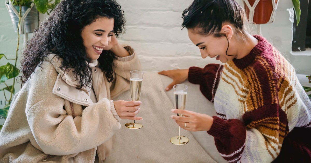 Young happy Hispanic female friends with glasses of champagne having fun sitting on sofa and celebrating holiday