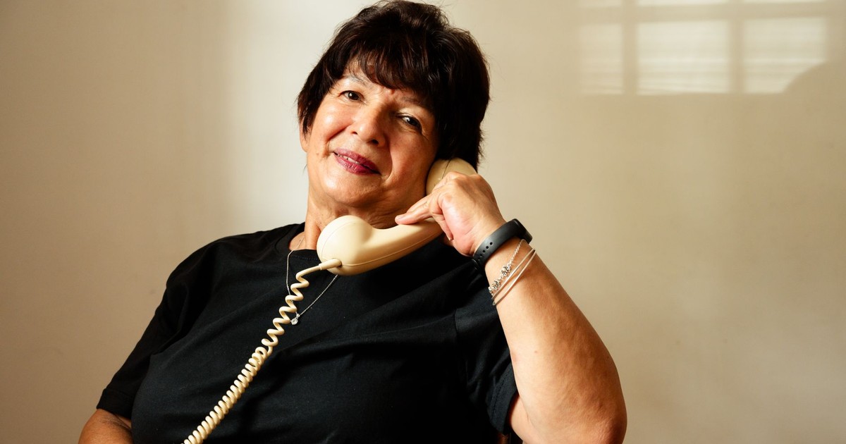 A senior woman wearing a black shirt smiles while talking on a vintage corded telephone indoors.