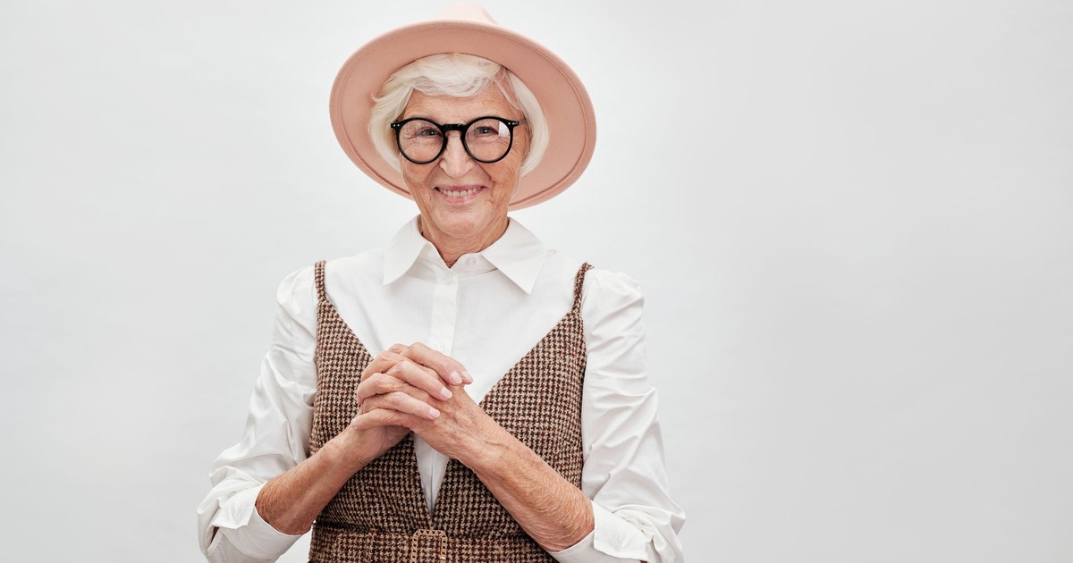 Fashionable senior woman smiling confidently in a stylish outfit with hat and eyeglasses.