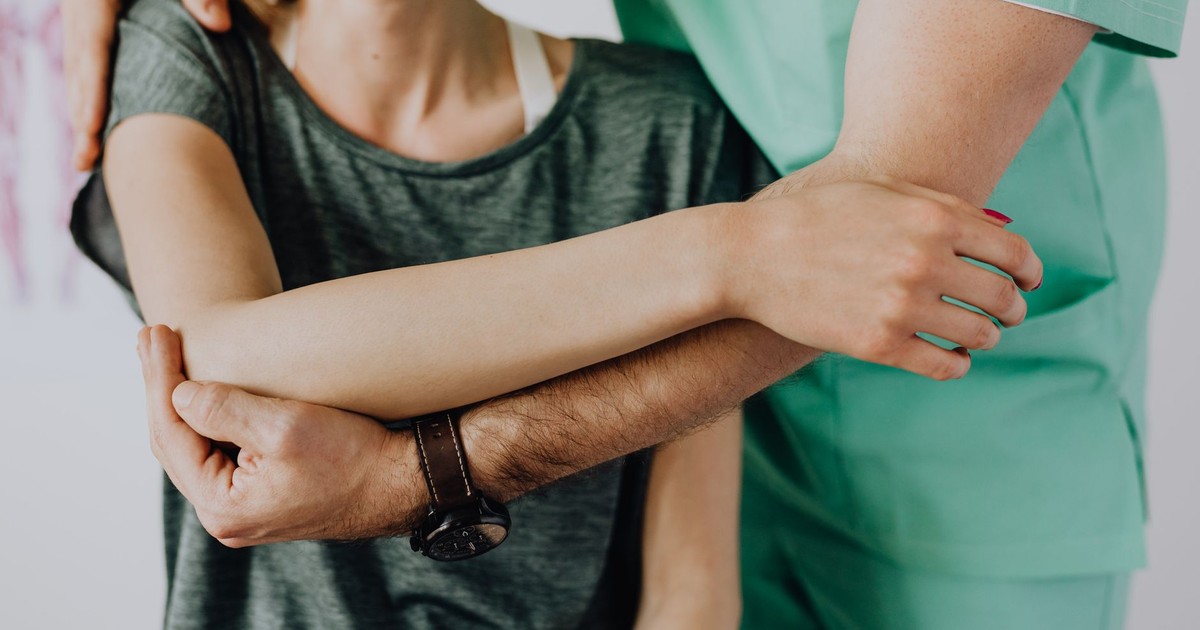 Crop anonymous chiropractor in uniform and wristwatch examining shoulder of smiling faceless woman in casual clothes in doctor office in clinic
