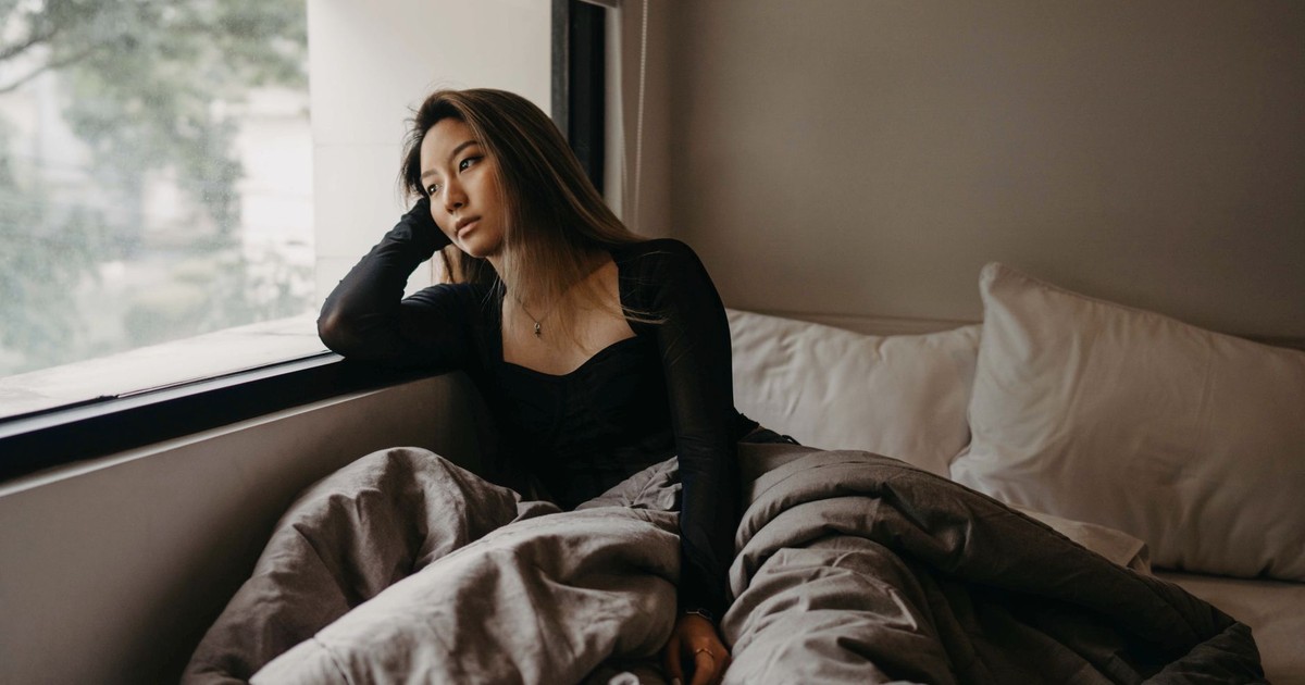 A young woman in a cozy room, pondering by the window, wrapped in blankets.