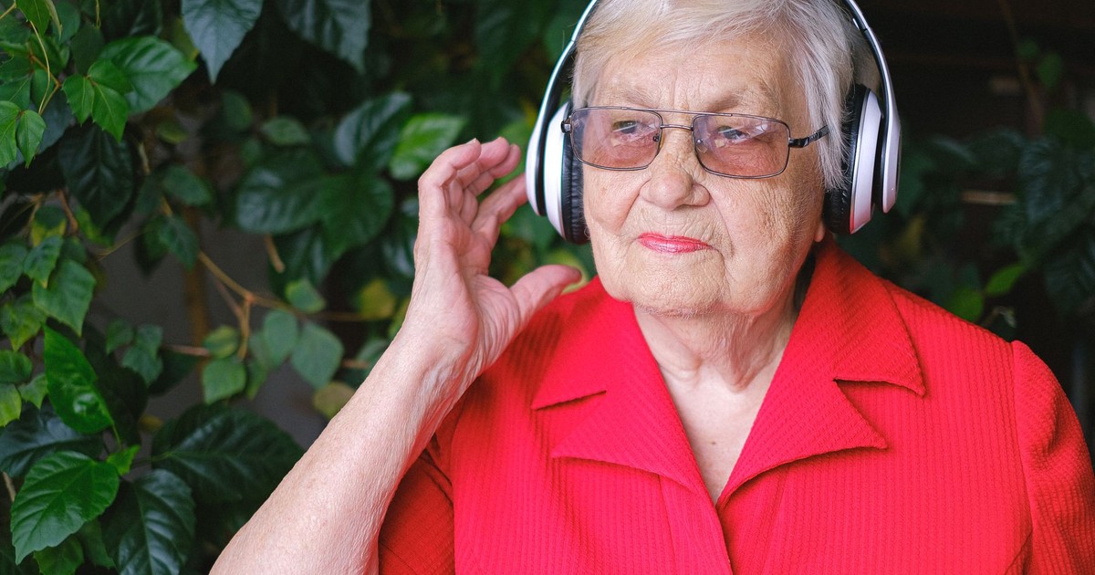 Senior thoughtful female in eyeglasses and wireless headphones listening to music near green leaves