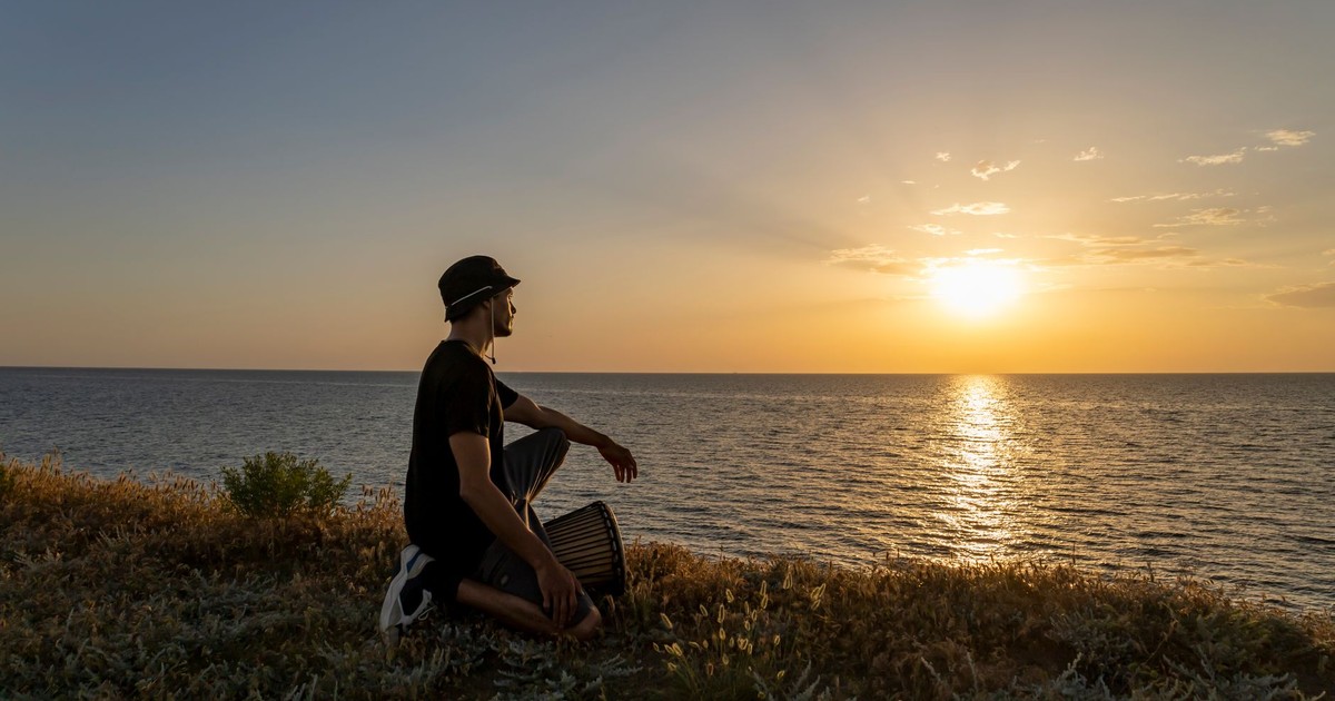 A man kneels on grassy cliff, playing drum, overlooking ocean sunset. Peaceful and serene moment.