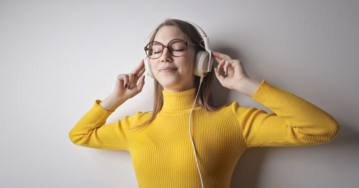 Woman in yellow sweater smiles while listening to music with headphones, eyes closed.