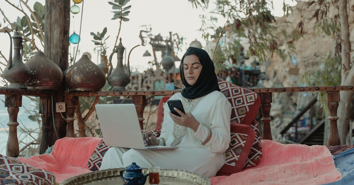 A woman in a hijab working on a laptop and phone in a cozy outdoor setting.