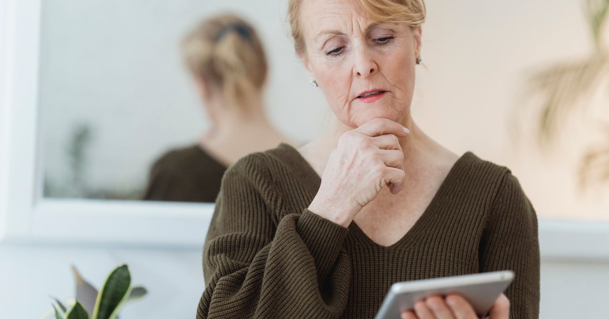 Thoughtful elderly female with mobile phone touching chin while reflecting in mirror in house room