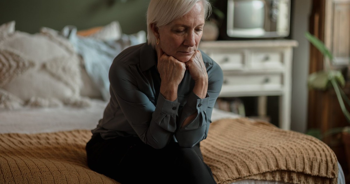 An elderly woman sitting on a bed, deep in thought, with a somber expression in a cozy bedroom.