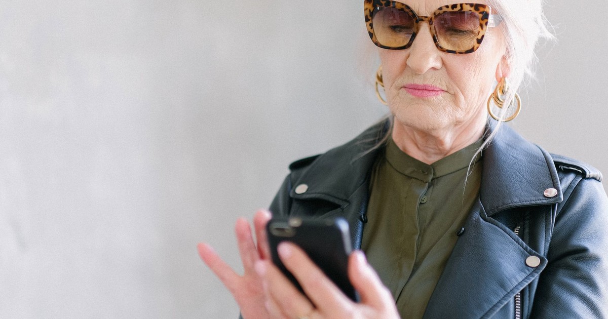 Crop elderly pensive female in fashionable outfit using mobile phone while texting on gray background