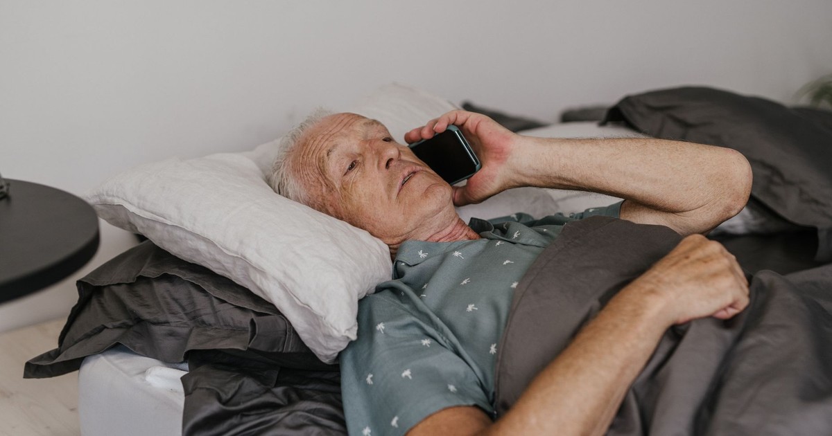 Senior man lying in bed with a smartphone, talking on a call, showcasing modern technology use.