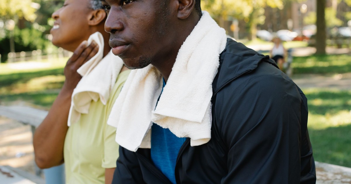 Two adults relaxing on a park bench with towels after a workout under the summer sun.