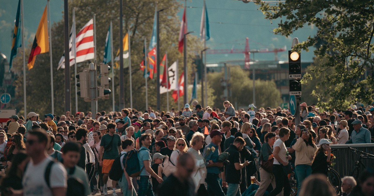 A bustling city street with a diverse crowd and international flags under a sunny sky.