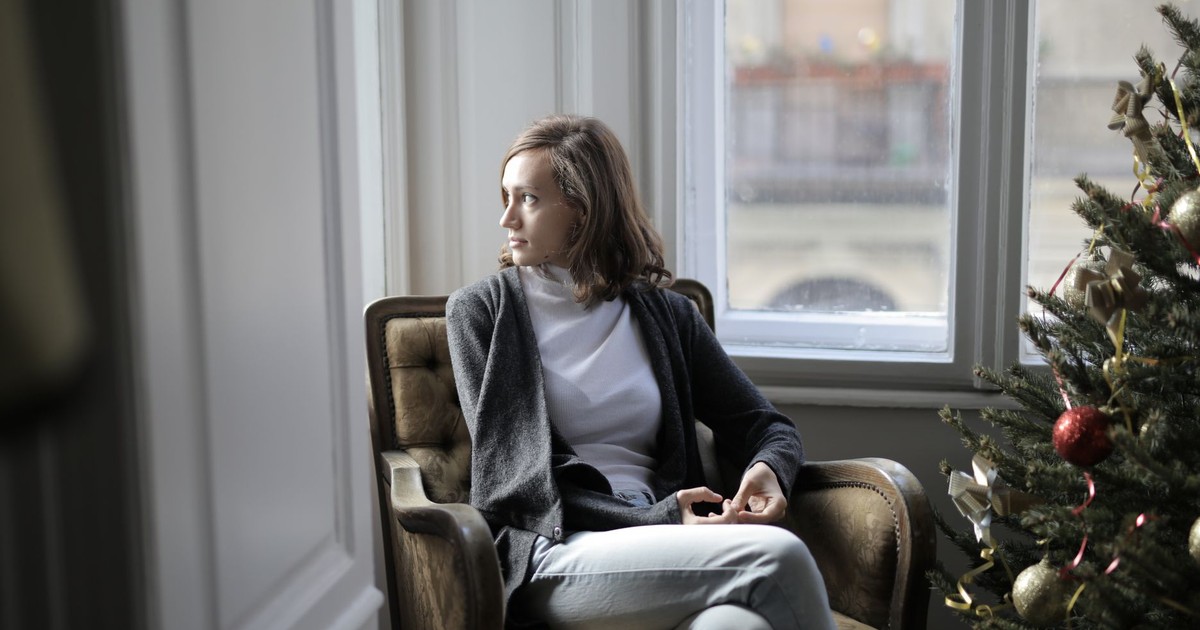Woman sitting in an armchair by a Christmas tree, gazing out the window with a contemplative look.
