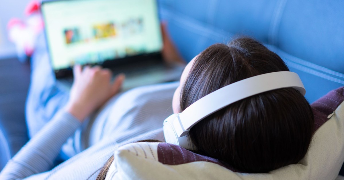 A woman lying on a sofa, enjoying music with headphones and browsing the internet on a laptop.