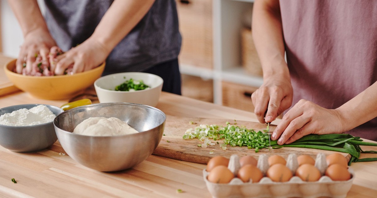 Two people preparing food with fresh ingredients on a wooden table in a cozy kitchen environment.