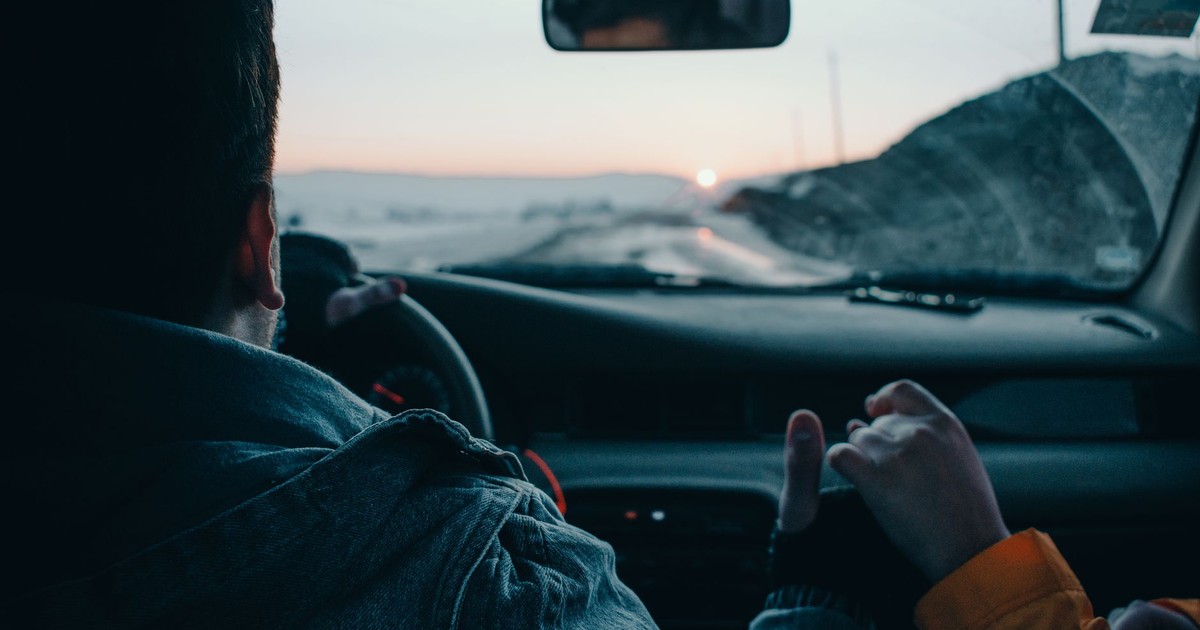 Back view of crop anonymous couple of travelers driving modern automobile along asphalt road at sunset