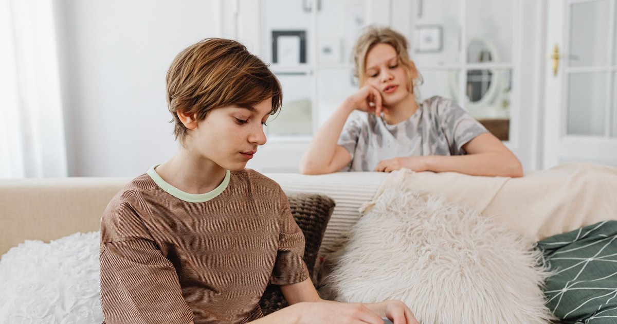 Two teenagers sitting on a comfortable sofa, one holding a bowl, in a well-lit living room.
