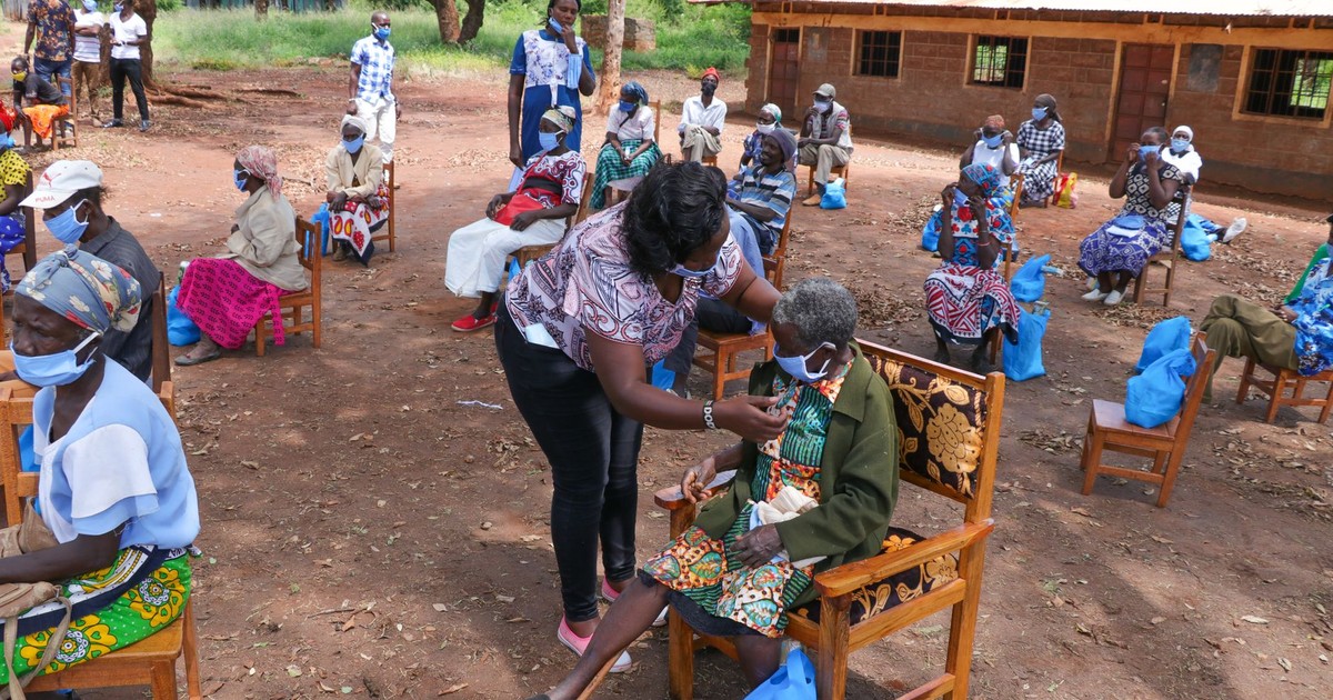 High angle of anonymous African female taking care of senior people in face masks sitting on chair near shabby building of nursing home in village