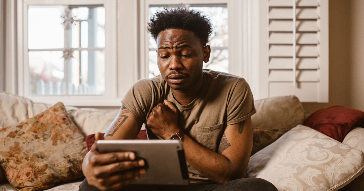 A man sitting on a sofa at home using a tablet with an emotional expression.